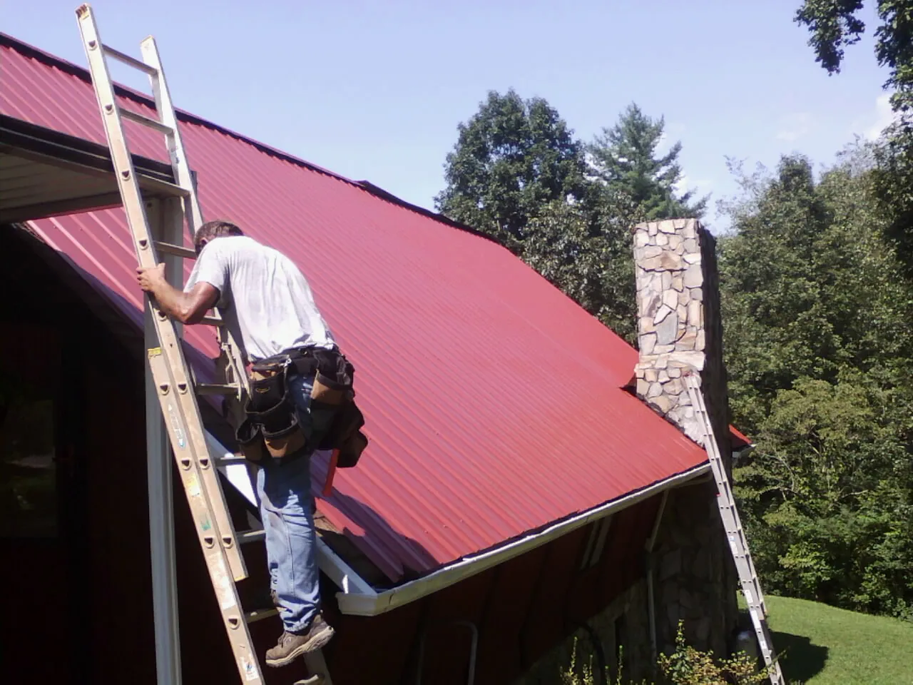 Roofer on a ladder beside a red metal roof and stone chimney — Soffit & Fascia Repair by Roofing Brothers in Yates City